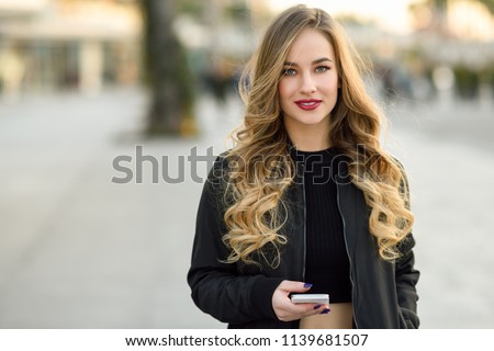 Blonde woman texting with her smart phone in urban background. Beautiful young girl wearing black jacket walking in the street. Pretty russian female with long wavy hair hairstyle.