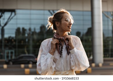 Blonde Woman Touches Her Silk Brown Scarf. Attractive Young Girl In Eyeglasses And White Blouse Smiles Outside.