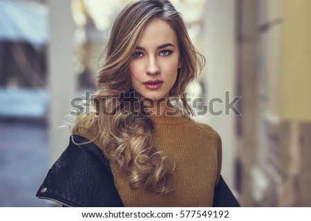 Blonde woman in urban background. Beautiful young girl wearing black leather jacket and mini skirt standing in the street. Pretty russian female with long wavy hair hairstyle and blue eyes.
