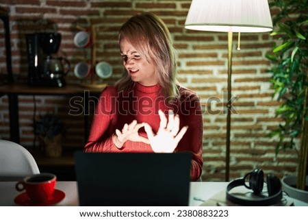 Blonde woman using laptop at night at home disgusted expression, displeased and fearful doing disgust face because aversion reaction. with hands raised 
