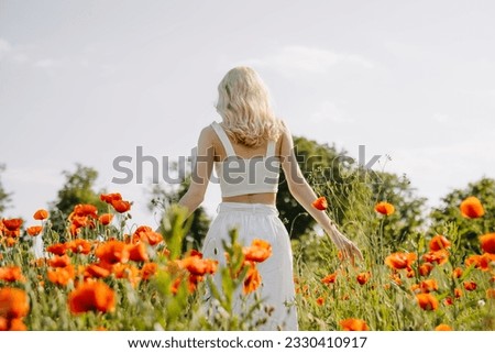Blonde woman walking in a field with wild red poppies, wearing a white dress.
