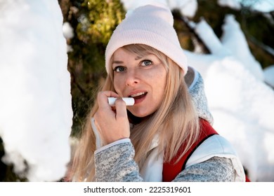 Blonde Woman with warm clothes applying lip balm on cold winter day outdoor. Female portrait close up putting cosmetic protection for lips on snowy season,. Copy space