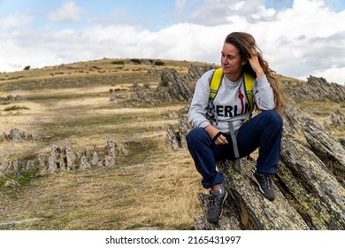 Blonde Woman With Wavy Hair, Sitting On A Rock While Resting On A Mountain Hiking Trail.