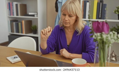 A blonde woman wearing a purple sweater attentively uses a laptop in a cozy home office with a coffee cup and a notebook on the table. - Powered by Shutterstock