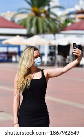 Blonde Woman Wearing A Surgical Mask And A Tank Top Taking A Photo Of Herself Using Her Phone On An Out Of Focus Background. Safety And Technology.