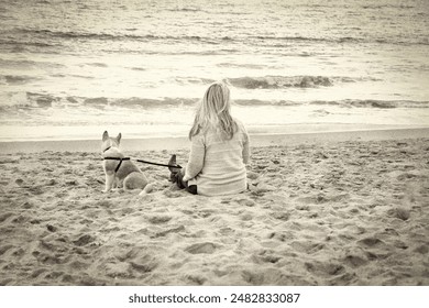 A blonde woman wearing a sweater sitting on the sand at a beach with her dog - Powered by Shutterstock