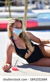 Blonde Woman Wearing A Swimsuit And A Surgical Mask Lying Down Next To A Cold Drink On An Out Of Focus Background. New Holidays And Safety Concept.