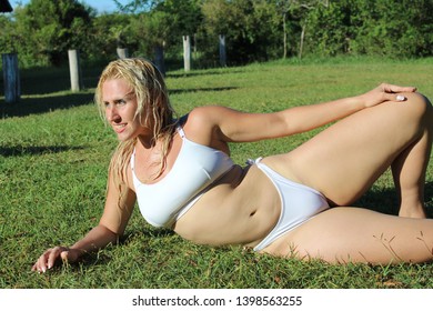 Blonde woman wearing a white bikini posing in the grass while sunbathing staring at the sun with green trees in the backgroud with her hair wet