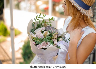 blonde woman in white dress and straw hat holds a bouquet of roses, eucalyptus and succulents packed in grey wrapping-paper