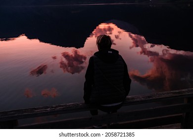 blonde woman in white outfit sitting back in front of the lake with red sunset sky reflection pink clouds romantic 