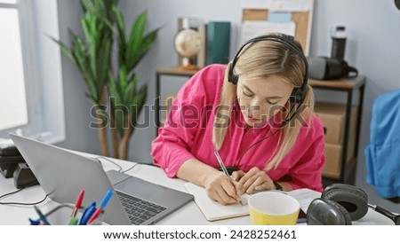 Blonde woman working indoors at office with laptop and headphones, taking notes while focused.