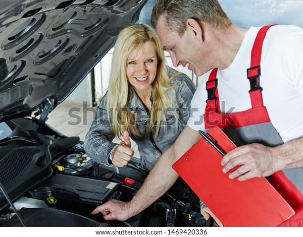 a\
blonde women stand with the mechanical master of the car service\
workshop at the open bonnet motor enginde of her car and looks\
satisfied and happy and show thumb up for this to\
him
