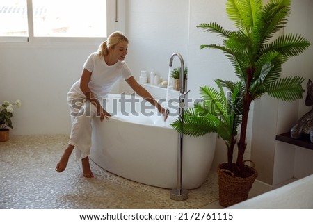 Blonde young beautiful female preparing warm bath with bubbles and pouring water.