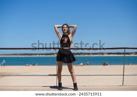 Blonde, young and beautiful woman dressed in black skirt and black top is on the promenade of the sea. The girl puts her hands behind her head. In the background the blue sea