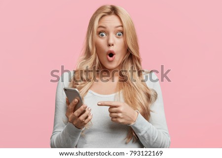 Blonde young female with pleasant appearance looks with terrified expression in smart phone, reads shocking news on webpage, isolated over pink background. Woman indicates at digital telephone
