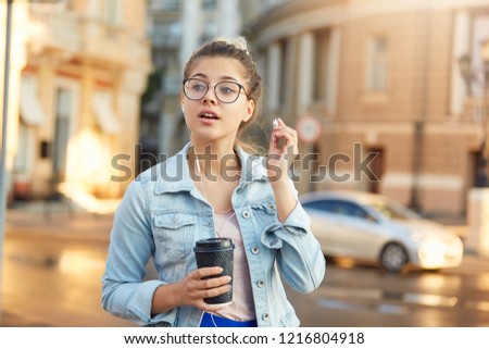 Blonde young girl in glasses walks around the city dressed casually, drinks coffee, listens in headphones to favorite music, took one earphone out of ear and turned around at the call of a passerby.
