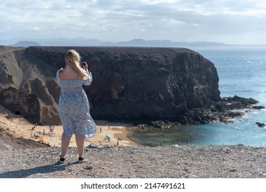 Blonde Young Girl with her back turned taking a photo with her phone to papagayo beach on Lanzarote canary islands. Travelers lifestyle and natural resources, beach of light sand and turquoise sea