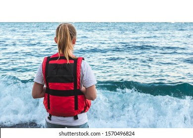 Blonde young girl in life jacket stands on the beach and watching waves. Lifeguard woman coast guard in bright red life vest looks in open sea. Ocean Shore Security Concept. Rough seas. View from back - Shutterstock ID 1570496824
