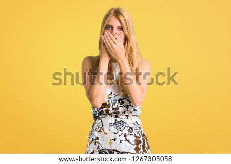 Blonde young girl in a summer dress covering mouth with both hands for saying something inappropriate. Can not speak on yellow background