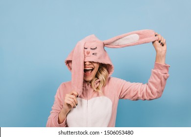 Blonde young lady posing in pink bunny costume. Studio shot of girl in kigurumi fooling around on blue background.