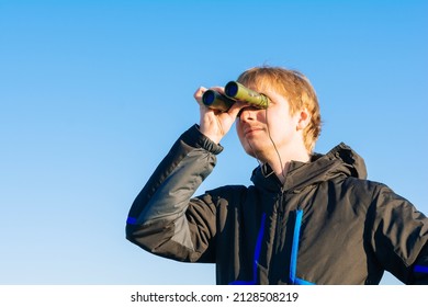 Blonde Young Man Looking Through Binoculars While Outdoors In Nature. Guy Looking Through Binoculars On The Blue Sky. Travel And Wildlife Concept