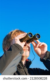 Blonde Young Man Looking Through Binoculars While Outdoors In Nature. Guy Looking Through Binoculars On The Blue Sky. Travel And Wildlife Concept