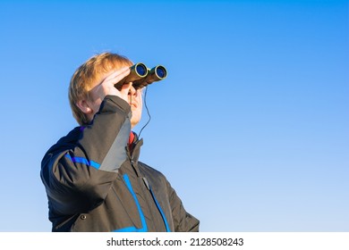 Blonde Young Man Looking Through Binoculars While Outdoors In Nature. Guy Looking Through Binoculars On The Blue Sky. Travel And Wildlife Concept