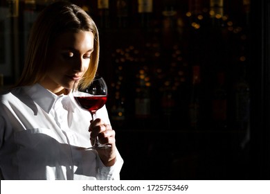 Blonde Young Pretty Girl Sommelier Holding And Smelling A Glass Of Red Wine. Appreciates The Aroma Of Wine.