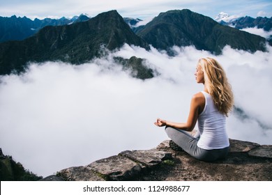 Blonde young woman with blonde hair doing meditation sitting in lotus on edge of rocky hill enjoying relax time of travel.Female tourist doing yoga from high peak mountain enjoying calm of nature - Powered by Shutterstock
