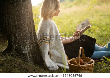 A blonde young woman sits on the grass under a tree with a basket of mushrooms and reads a book. Girl resting on a small picnic near the forest