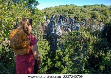 A blonde young woman with a straw hat on her back looking at Palanquinhos Canyon, in Caxias do Sul, Rio Grande do Sul, Brazil