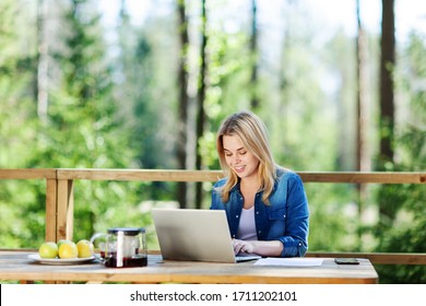 Blonde young woman working on laptop computer sitting at table on wooden balcony of country house - Powered by Shutterstock