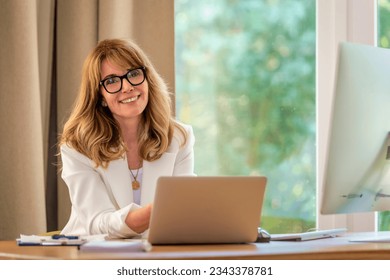 Blondehaired businesswoman sittint at her desk and working on her laptop. Professional female wearing eyewear and white blazer.