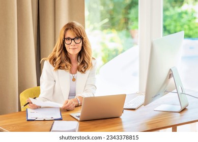 Blondehaired businesswoman sittint at her desk and working on her laptop. Professional female wearing eyewear and white blazer.
