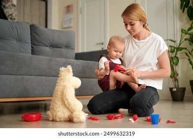 Blonde-haired mum and baby girl of one and a half years old playing hospital with toy first aid kit at home on floor, mum taps girl's knee with toy hammer. - Powered by Shutterstock