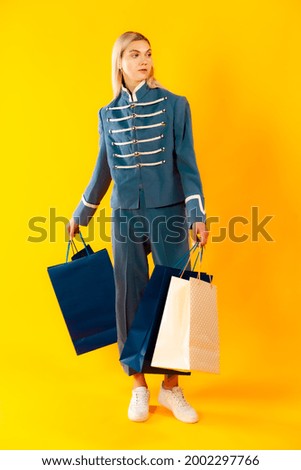 Blondie in military uniform looking aside ready to go isolated on yellow. Blonde hair girl posing with shopping paper bags. Shopping and presenting concept. Photo for fashion, beauty, art and adS
