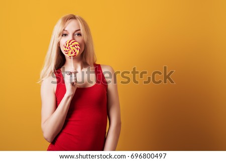 Blondy girl holding big candy looking at camera isolated over the yellow background.