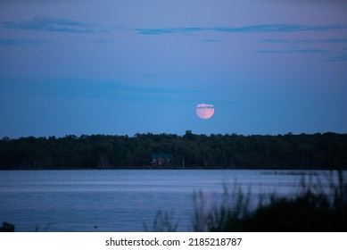 Blood Moon Perched Above Tree-line Over Canada And Waters That Are Lake Huron. Rustic Lake House On Shore.