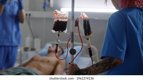 Blood Transfusion To Young Male Patient In Hospital Room. Nurse Pushing Iv Set With Blood For Unconscious Man Lying In Hospital Bed In Intensive Care Unit