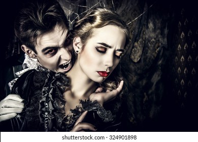 Bloodthirsty male vampire in medieval dress is biting a beautiful lady. Halloween. 