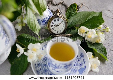 Blooming delicate jasmine flowers and a cup of tonic green tea decorated with a mechanical clock on a chain.