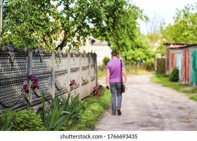 Blooming Rural Street In The Summer. The Delivery Worker Brought The Order To The Specified Address. A Guy With A Shoulder Bag Is Going On The Village Road. Delivery, Quarantine. Defocused