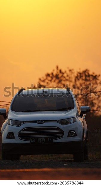 Blora,\
Indonesia - August 25, 2021 :  White compact SUV Ford Ecosport with\
sport and modern design parked at sunset. Automotive industry. Car\
maintenance business background. Beautiful\
sky.