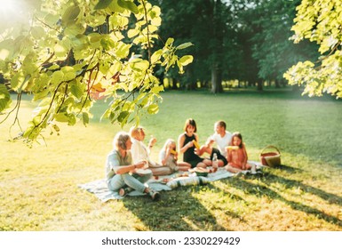 Blossom linden tree branch with Big family sitting on the picnic blanket in city park during weekend Sunday sunny day. They smiling and laughing and eating lunch. Nature outdoors activities concept. - Powered by Shutterstock