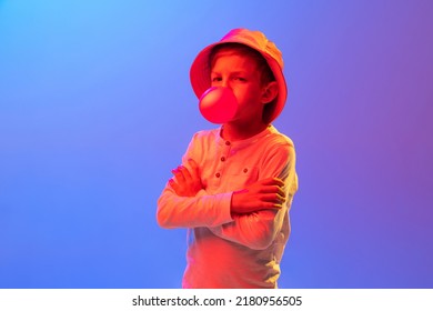 Blowing Bubble Gum. Studio Shot Of Stylish Little Boy, Kid Posing Isolated On Gradient Blue-purple Background In Neon Light. Concept Of Emotions, Facial Expression, Fashion, Hobbies, Active Lifestyle