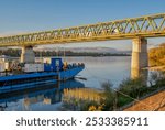 A blue barge docked beneath the Ujpest railway bridge on the Danube, with a passing train above. The autumn sunlight casts a warm glow on the serene river scene.