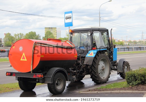 Blue Belarus tractor watering machine street cleaner\
with orange barrel pours asphalted  sidewalk back view, street\
washing in Moscow, city improvement by municipal services on a\
sunny summer day