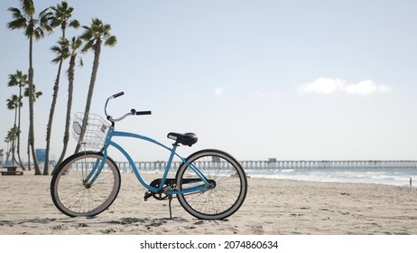 Blue Bicycle, Cruiser Bike By Sandy Ocean Beach, Pacific Coast, Oceanside Pier California USA. Summertime Vacations, Sea Shore. Vintage Cycle, Palms, Sky, Lifeguard Tower Watchtower Hut, Car Truck.