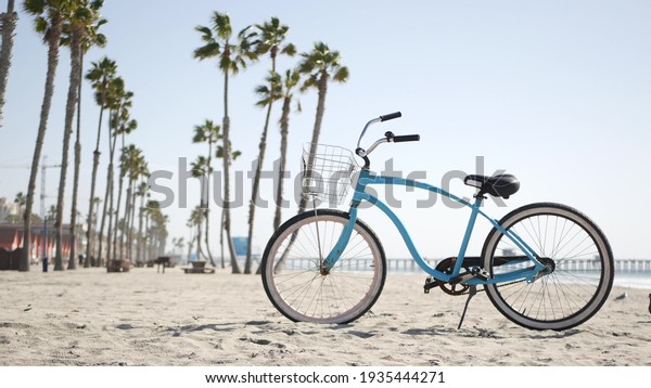 Blue bicycle, cruiser bike by sandy ocean beach,\
pacific coast near Oceanside pier, California USA. Summertime\
vacations, sea shore. Vintage cycle, tropical palm trees, lifeguard\
tower watchtower hut