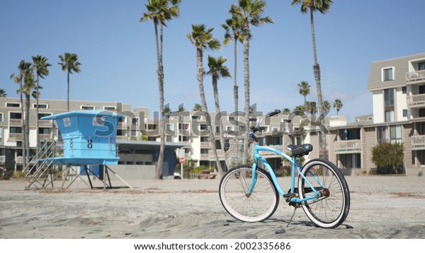 Blue bicycle, cruiser bike by ocean beach, pacific\
coast, Oceanside California USA. Summertime vacations, sea shore.\
Vintage cycle by beachfront houses, waterfront lodging. Lifeguard\
and palm trees.